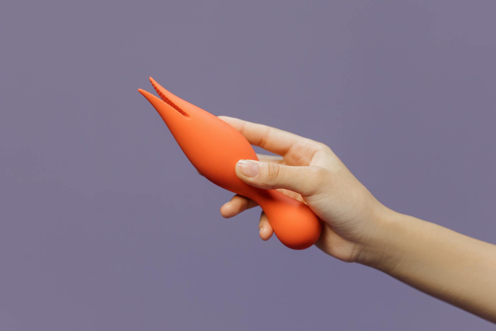 Mano sosteniendo un vibrador naranja moderno contra un fondo minimalista morado, mostrando sensualidad contemporánea.
